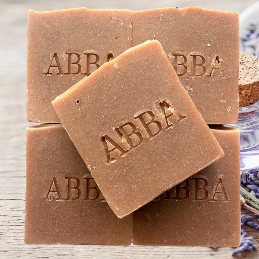 Cuatro barras cuadradas de jabón artesanal de la marca ABBA apiladas sobre una superficie rústica de madera, con una pastilla colocada en diagonal en la parte superior. El jabón es de un color marrón medio cálido y lleva el nombre de la marca «ABBA» grabado en relieve en la parte frontal.