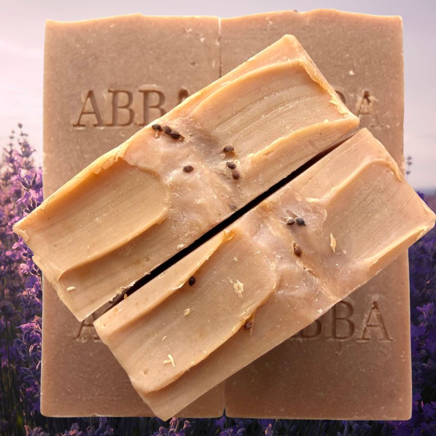 Dos barras rectangulares de jabón artesanal de color beige cremoso con pequeñas inclusiones botánicas oscuras, dispuestas en diagonal frente a una pastilla más grande del mismo color. El jabón se fotografía con un fondo de un suave campo de lavanda.
