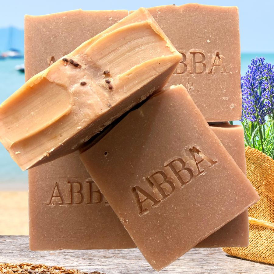 Tres barras de jabón hecho a mano ABBA apiladas sobre una superficie de madera con un fondo de playa y campo de lavanda. El jabón es de un marrón medio cálido con una capa de color canela más claro visible en la superficie cortada y la marca 'ABB A' está grabada en cada barra.