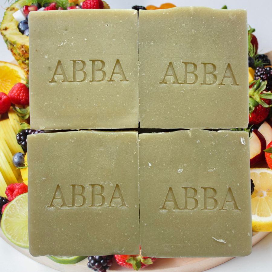 Cuatro barras rectangulares de jabón de la marca ABBA dispuestas en una cuadrícula de 2x2, con el nombre de la marca grabado en cada una. El jabón es de un color verde oliva suave y se presenta en un plato rodeado de frutas frescas como fresas, arándanos, naranjas y limas.