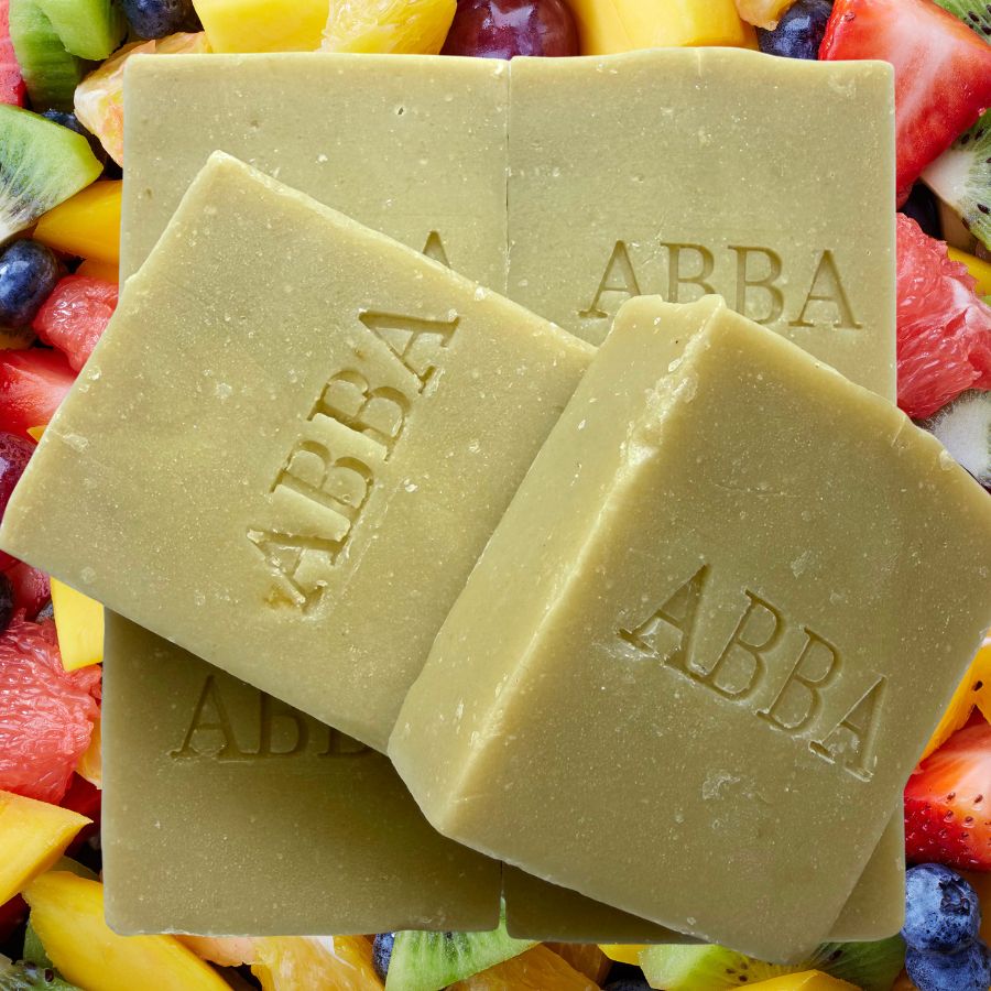 Cuatro barras de jabón artesanal ABBA de color verde oliva apiladas una encima de la otra, con el nombre de la marca 'ABBA' grabado en relieve en un lateral, sobre un colorido fondo de frutas en rodajas que incluyen sandía, mango, kiwi y arándanos.