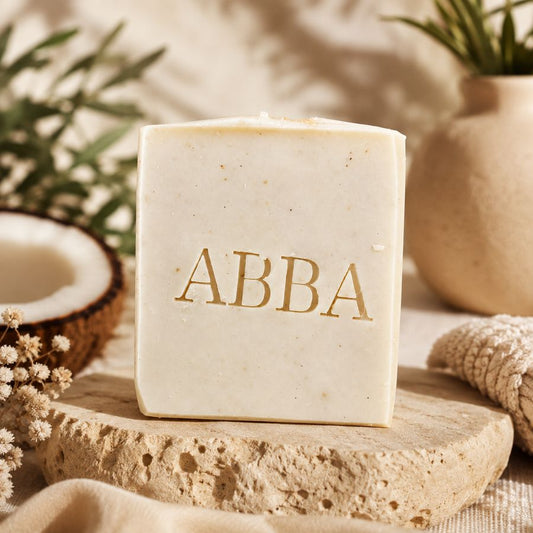 Una barra rectangular de jabón color crema con el nombre 'ABBA' impreso en letras doradas, expuesta sobre una losa de piedra texturizada con medio coco, flores secas y una maceta beige al fondo.