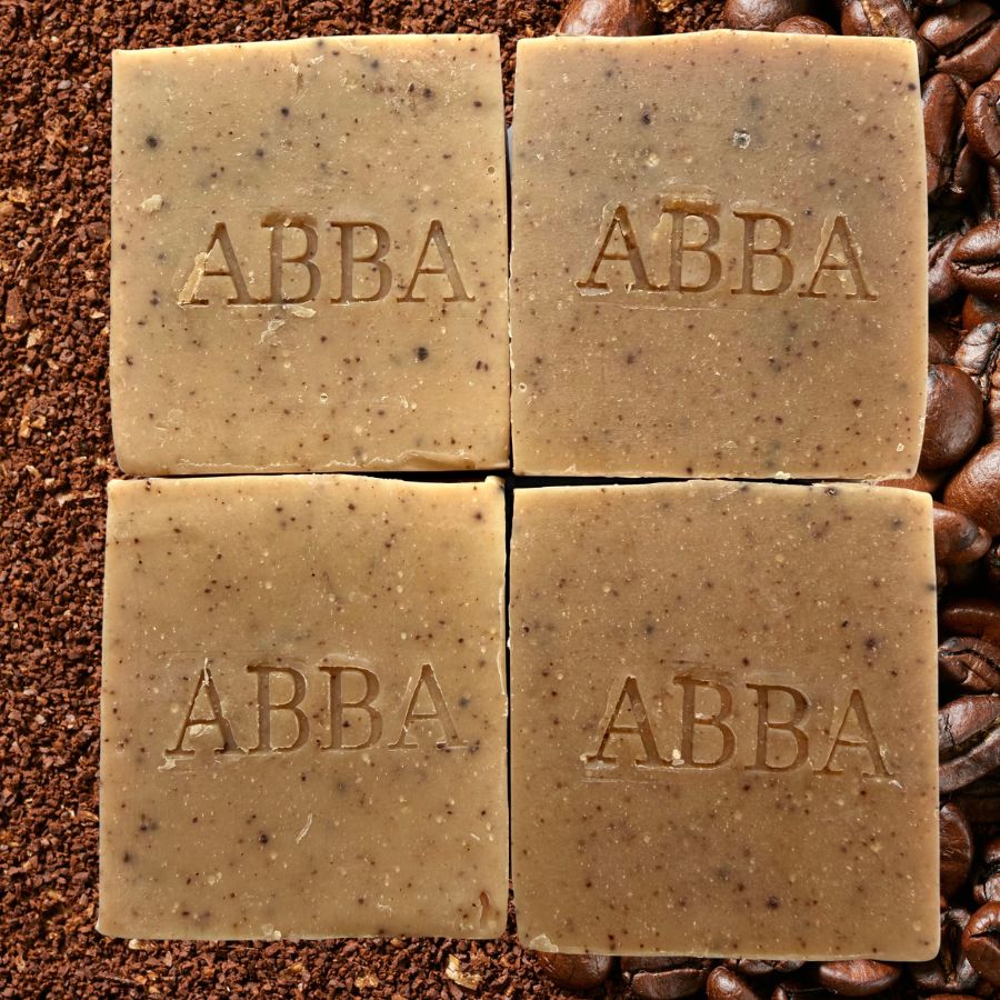 Cuatro barras cuadradas de jabón artesanal ABBA dispuestas en una cuadrícula de 2x2, con la inscripción "ABBA" grabada en cada una. El jabón es de color marrón claro con motas oscuras y está fotografiado sobre un fondo de granos y posos de café.