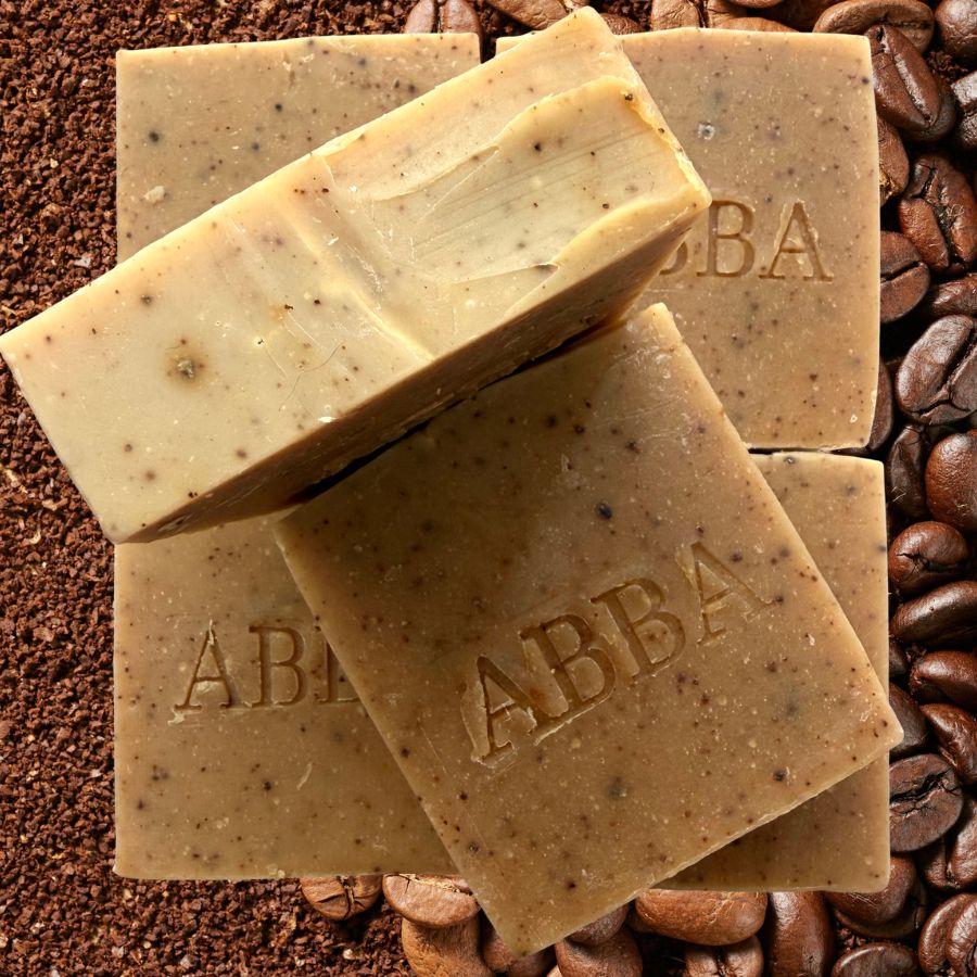 Tres barras rectangulares de jabón de la marca ABBA con motas de café, apiladas una encima de la otra sobre un fondo de granos y posos de café.