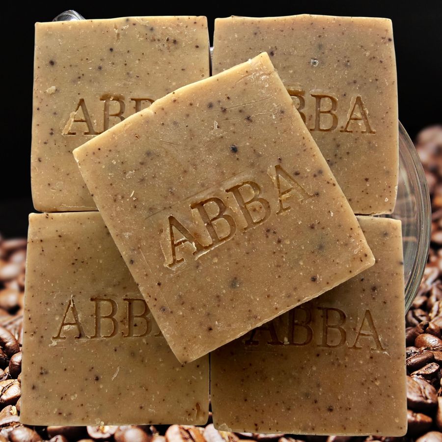 Cuatro barras cuadradas de jabón de la marca ABBA apiladas, con la pastilla central inclinada para mostrar el nombre de la marca grabado en su superficie. El jabón tiene un color beige cálido con motas marrón oscuro, y las pastillas están dispuestas sobre una base de granos de café sobre un fondo oscuro.