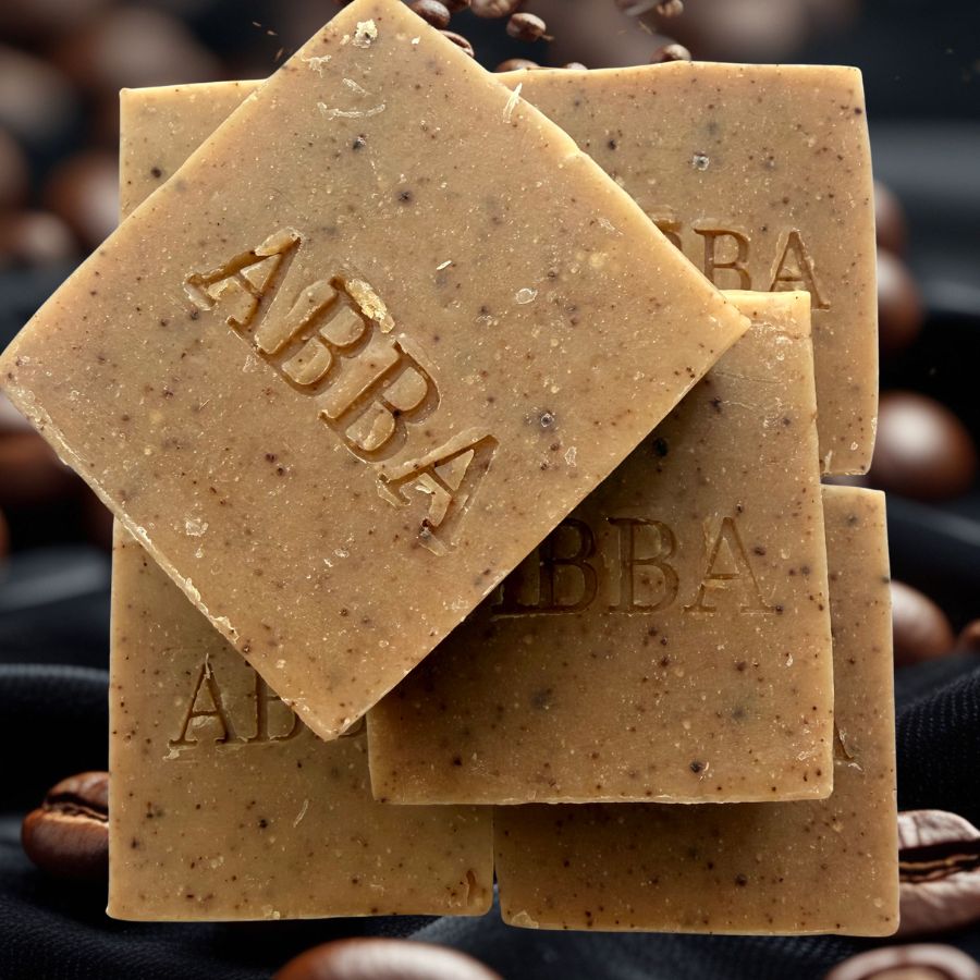 Cuatro barras cuadradas de jabón de la marca ABBA apiladas, de un cálido color marrón tostado con motas oscuras que recuerdan a granos de café y la inscripción "ABBA" en relieve, sobre un fondo oscuro con granos de café dispersos.