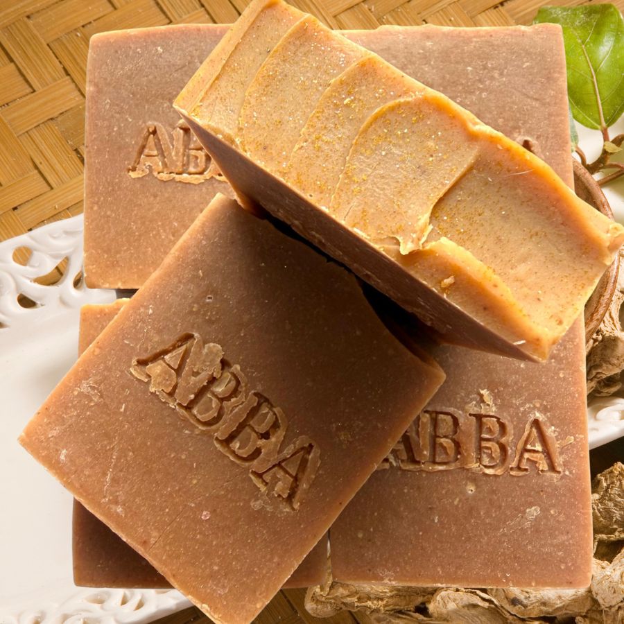 Cuatro barras rectangulares de jabón artesanal de la marca ABBA apiladas sobre un plato blanco, una de ellas rota para mostrar su textura en capas. Los jabones son de un tono marrón medio cálido con una capa superior de color marrón dorado más claro, sobre un fondo de raíz de jengibre y una estera de bambú.