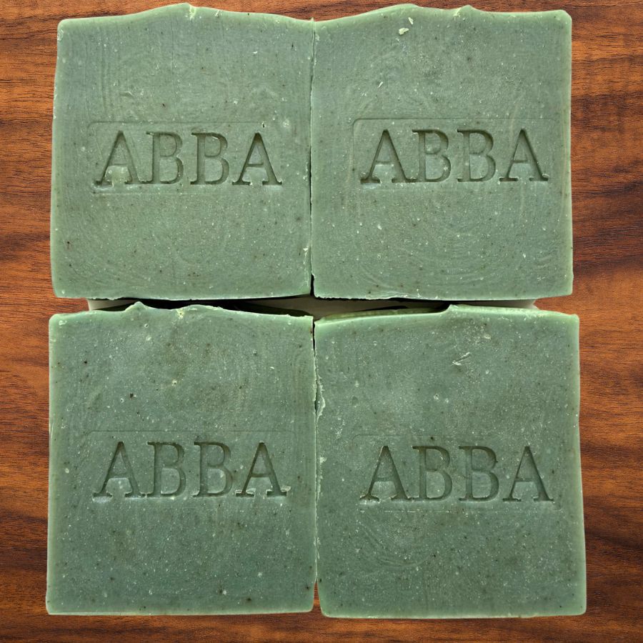 Cuatro barras rectangulares de jabón de la marca ABBA, de un suave color verde salvia, dispuestas en una cuadrícula de 2x2 sobre una superficie de madera marrón oscuro. El nombre de la marca, «ABBA», está grabado en relieve en el centro de cada pastilla.