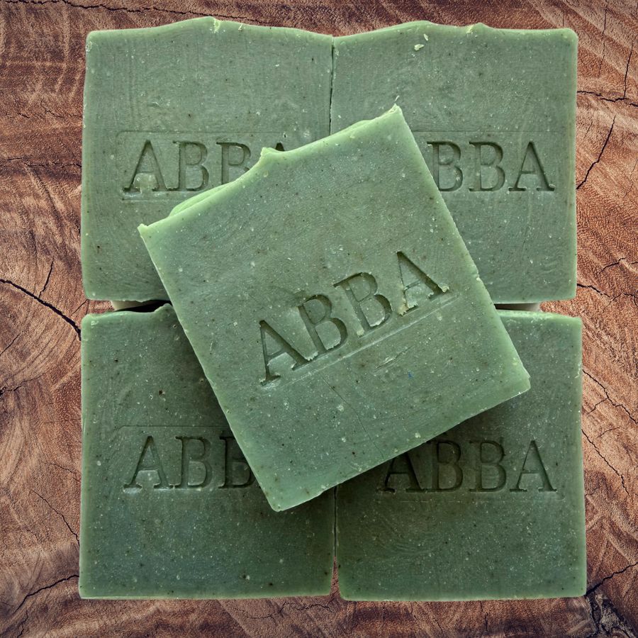 Cuatro barras rectangulares de jabón verde oliva dispuestas sobre una superficie de madera rústica, con una pastilla encima. El jabón lleva grabado el nombre de la marca «ABBA».